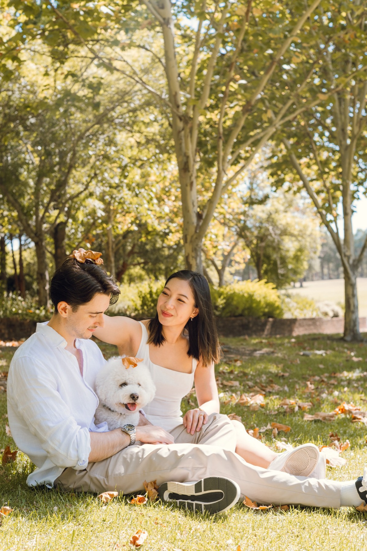 Damon and Christina sitting on the grass in a sunny park with their fluffy white dog, Marlo